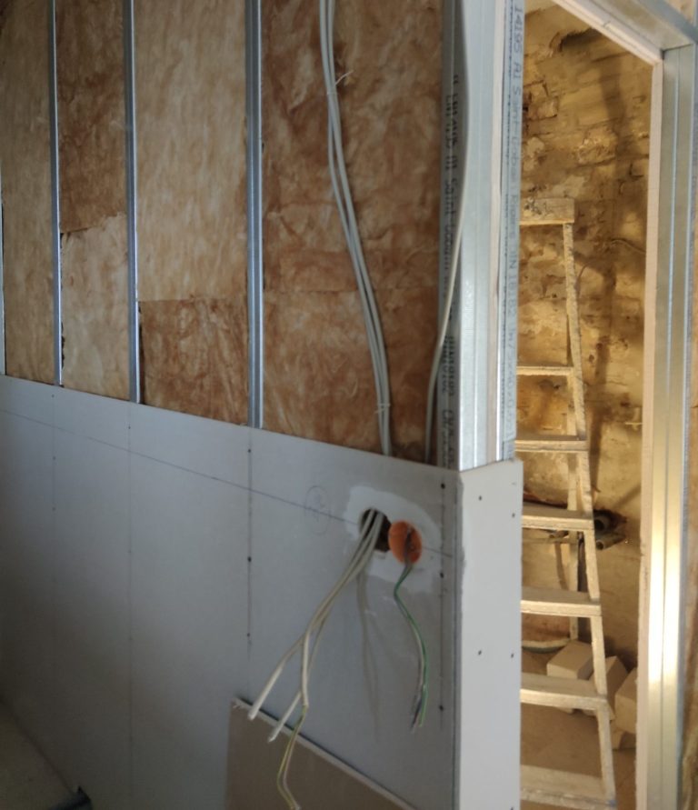 Unfertige Wand mit Steckdose und Leiter im Hintergrund.